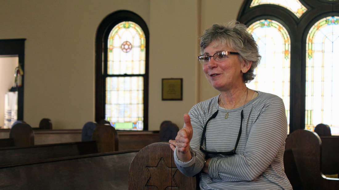 Gail Goldberg leans on a wooden pew in the sanctuary of Ahavath Rayim. Behind her are stained glass windows.