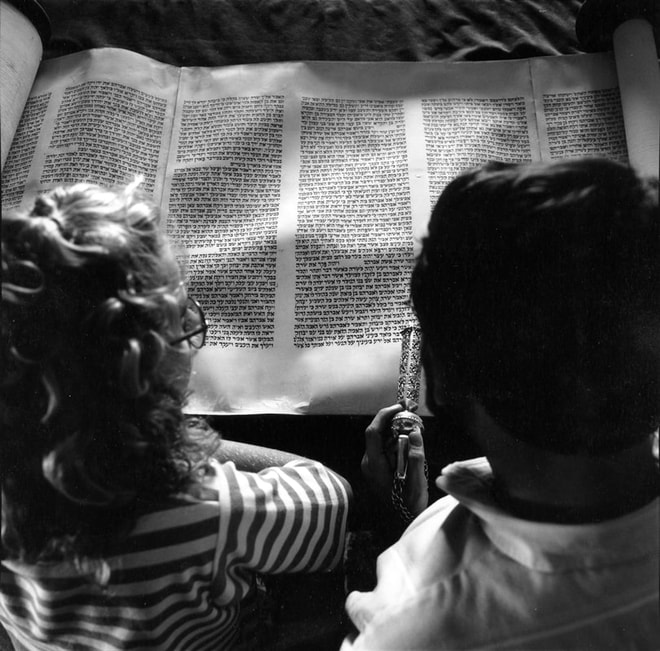 Black and white photo presents an overhead view of a man and a girl as they examine a Torah scroll together. He holds an ornate yad, the ritual tool used by readers to track their place in text without touching the Torah parchment themselves.