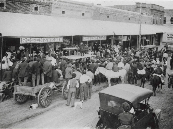 Crowded street scene including horses, wagons, cars, and pedestrians, as a young performer stands on a platform in front of Rosensweig's Store.