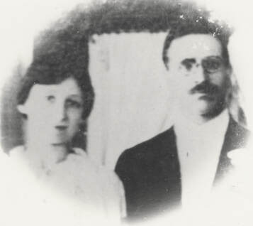 Slightly blurry photograph of a woman in a white dress and a man with mustache and glasses who is wearing a dark suit with a white shirt.