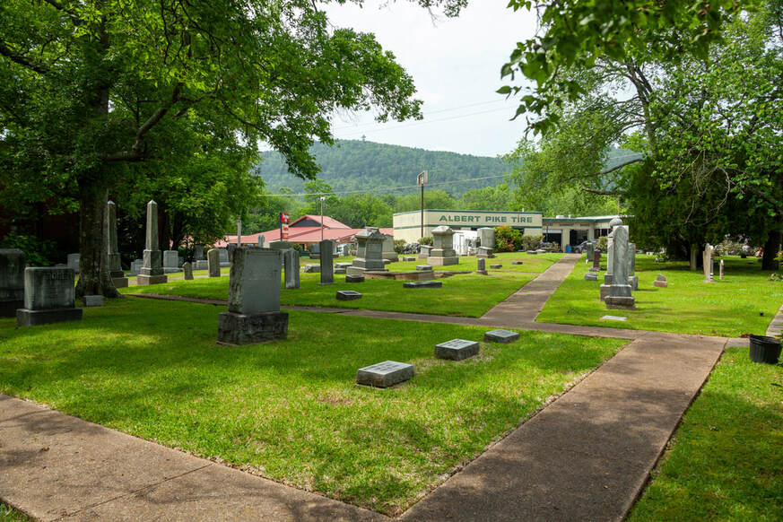 Green foliage frames a collection of headstones at Jewish Rest Cemetery. A tire shop and another business sit in the near background, with forested hills rising behind them.