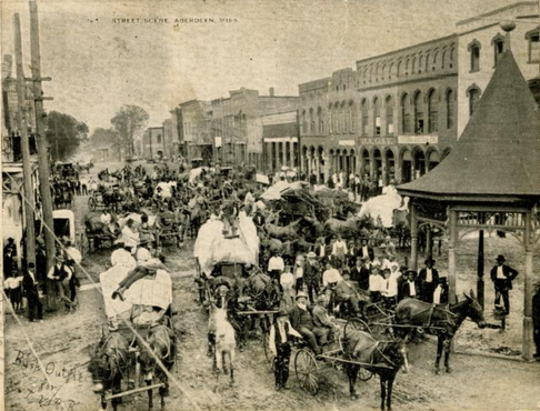 Black and white photo depicts a dirt street full of horse-drawn wagons. Two-story storefronts line the right side of the road, and there is a gazebo in the foreground. Standing figures include white and black men.