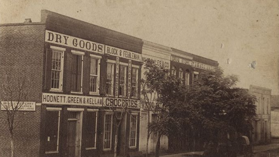 A row of storefronts bear names of proprietors: Hodnett, Green, and Kellam's dry goods store is in the foreground, with Block & Feibleman (dry goods, hardwards, and groceries) nextdoor. Three more storefronts are partially obscured by trees. A buggy is parked in front of the trees.