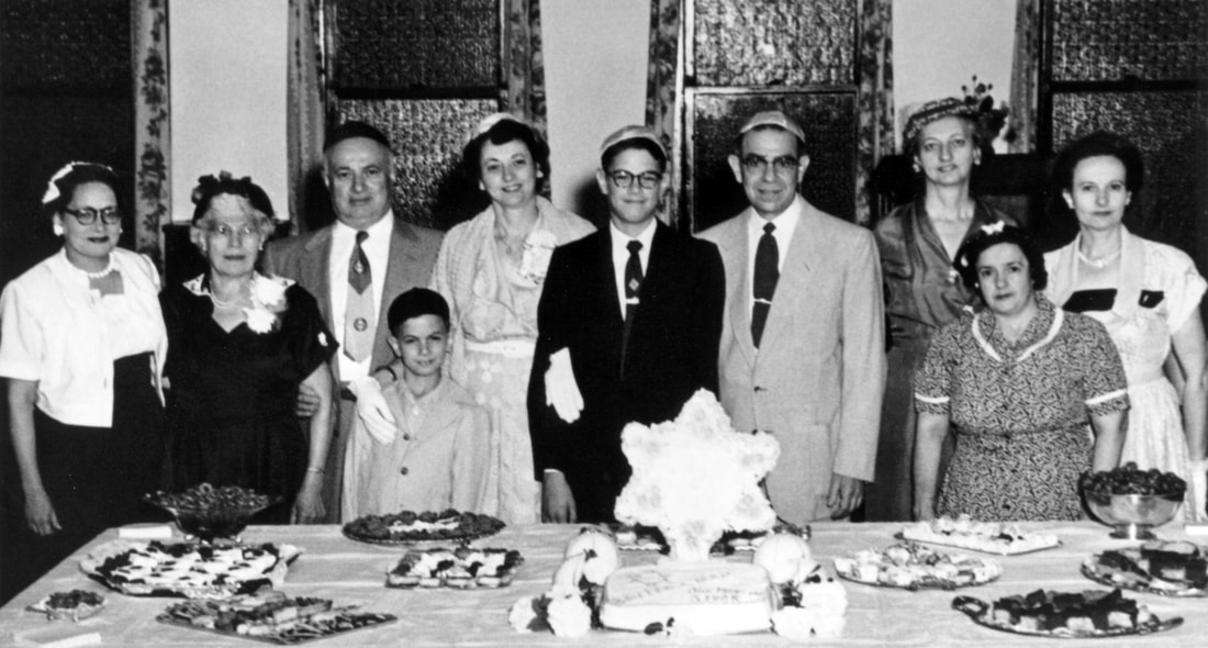 Black and white photo: a young teen in a dark suit stands in a row of formally dressed adults (and one child). In front of the group is a table set with food for a reception. In the center is a cake with a large Star of David rising from it.