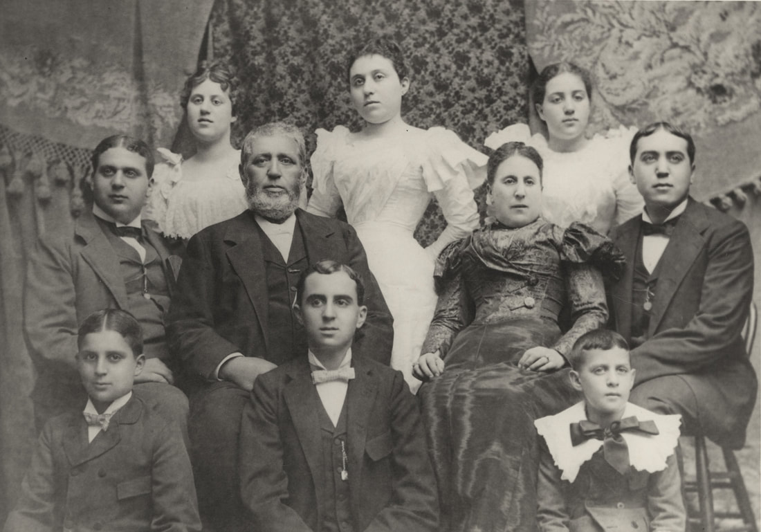 A family of ten sit for a photographic portrait. Parents Philip and Clara Pfeifer wear a three-piece suit and a dark dress. Three daughters in frilly white dresses stand behind them, and five sons wear dark suits, the youngest with a frilly white collar.