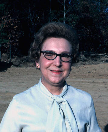 Celeste Orkin in white blouse and horn-rimmed glasses, standing in front of bare ground and trees.