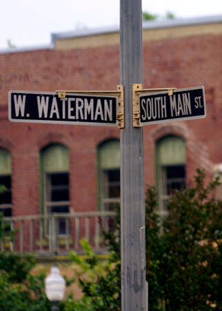 Signs for West Waterman and South Main Streets adorn a gray signpost. Trees and a downtown retail building sit in the background.